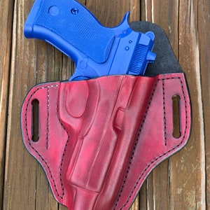 May include: A blue handgun rests in a red leather holster. The holster is stitched with black thread and has a molded design. The background is a wooden surface.