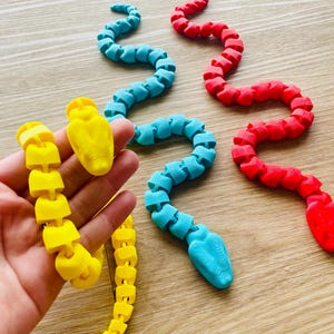 Könnte beinhalten: Drei flexible, segmentierte Schlangenspielzeuge in Gelb, Blau und Rot. Die Schlangen sind aus Kunststoff und haben ein realistisches Schlangenkopfdesign.