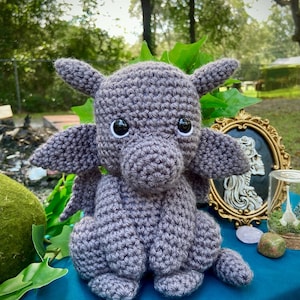 May include: A crocheted gray dragon with large black eyes. The dragon is sitting on a blue surface with green foliage in the background.