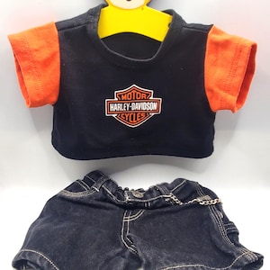 May include: A black t-shirt with orange sleeves and a Harley-Davidson Cycles logo, paired with dark denim shorts. The outfit is designed for a teddy bear, displayed on a yellow bear-shaped hanger.