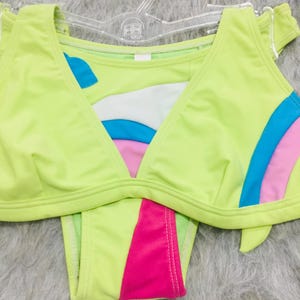 Puede incluir: Un conjunto de bikini verde lima con top triangular y braguita de talle bajo. El top presenta un diseño de cuello en V con detalles blancos, azules y rosas. La braguita tiene un panel rosa en la parte delantera. El traje de baño se muestra en una percha transparente.
