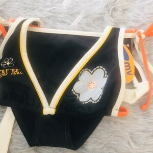 May include: Black bikini with white and yellow trim. The top has a floral applique and the bottom has a classic cut. The straps are orange and white. The bikini is displayed on a fluffy, light-coloured surface.