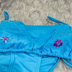 Peut inclure: Un ensemble de bikini turquoise avec un haut dos nu et un bas assorti. Le haut est orné de petits strass rouges et roses et d'un motif floral. Le bikini est présenté sur un cintre en plastique transparent.