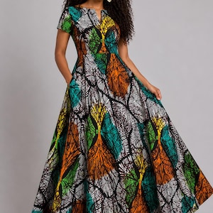 May include: A long, patterned dress featuring a leaf design in green, orange, yellow, and white on a black base. The dress has short sleeves, a V-neck, and pockets. The model is wearing a matching headband and gold earrings. The dress is floor-length.