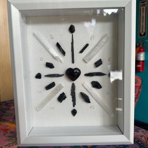 May include: A white framed shadow box with a black heart in the center surrounded by black and white crystals.
