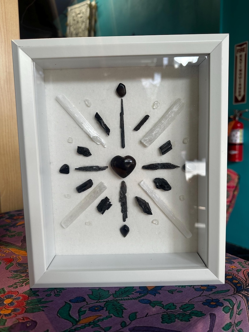 May include: A white framed shadow box with a black heart in the center surrounded by black and white crystals.