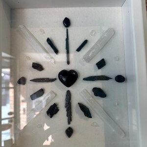 May include: A framed crystal grid with black and white crystals arranged in a starburst pattern. A black heart-shaped crystal is in the center of the grid.