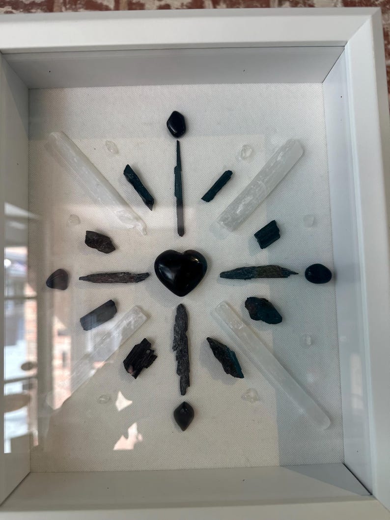May include: A framed crystal grid with black and white crystals arranged in a starburst pattern. A black heart-shaped crystal is in the center of the grid.