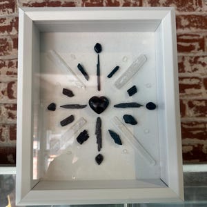 May include: A white framed shadow box with a black heart in the center surrounded by black and white crystals.