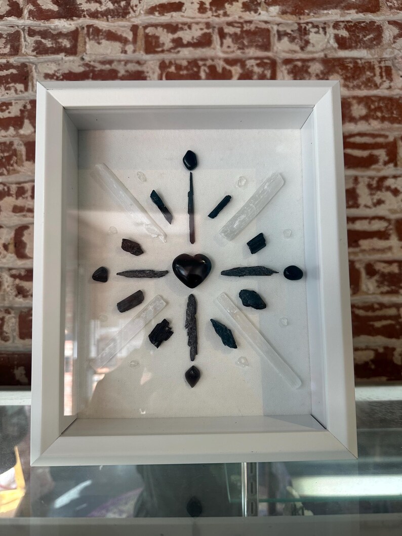 May include: A white framed shadow box with a black heart in the center surrounded by black and white crystals.