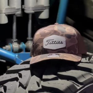 May include: A brown camouflage trucker hat with a white patch that reads "Titties" in a script font. The hat is resting on a large, black tire with deep treads. The hat has a mesh back.