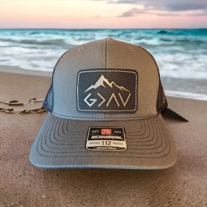 May include: A gray and black trucker hat with a black leather patch featuring a mountain range and the text "G>A>V". The hat has a black mesh back and a Richardson 112 label.