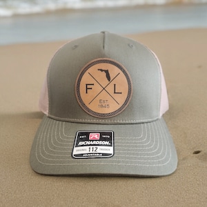 May include: A green and pink trucker hat with a brown leather patch featuring the letters "F" and "L" with a silhouette of the state of Florida in between. The patch also includes the text "Est. 1845". The hat is labeled "Richardson 112 Original Trucker Adjustable".