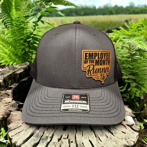 May include: A black and gray trucker hat with a brown leather patch that says "Employee of the Month Runner Up". The hat is adjustable and has a Richardson logo.