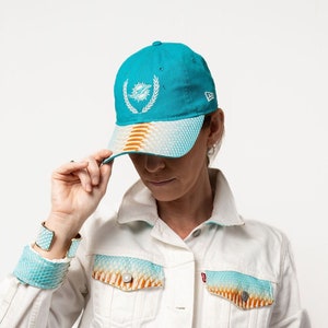 Może przedstawiać: Biała kurtka dżinsowa z akcentami w kolorze turkusowym i pomarańczowym w kształcie skóry węża na kieszeniach i mankietach. Kurtka zapinana jest na guziki i ma klasyczny styl dżinsowy. Modelka nosi turkusową i pomarańczową czapkę bejsbolową z białym logo delfina i napisem "New Era" z boku.