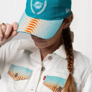 K&ouml;nnte beinhalten: Eine t&uuml;rkisfarbene Baseballkappe mit einem wei&szlig;en gestickten Miami Dolphins-Logo und einem wei&szlig;en, orangefarbenen und blauen gemusterten Schirm.