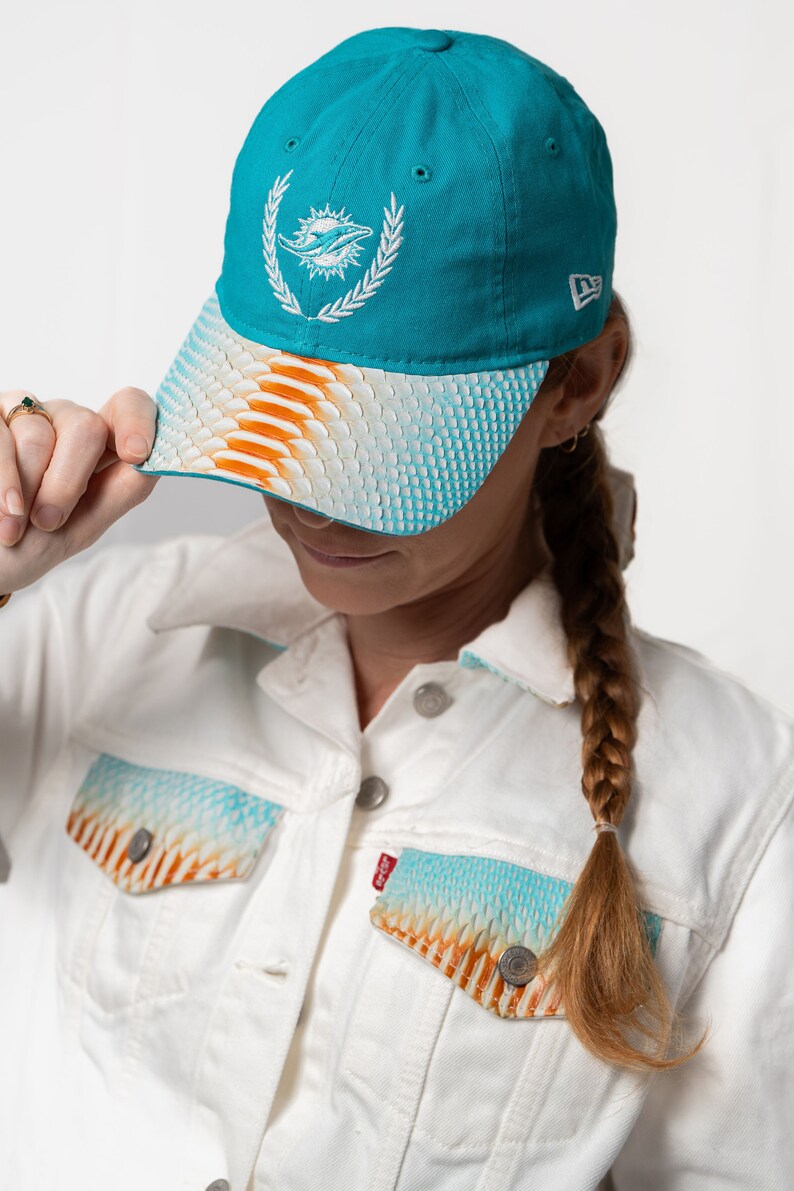 K&ouml;nnte beinhalten: Eine t&uuml;rkisfarbene Baseballkappe mit einem wei&szlig;en gestickten Miami Dolphins-Logo und einem wei&szlig;en, orangefarbenen und blauen gemusterten Schirm.