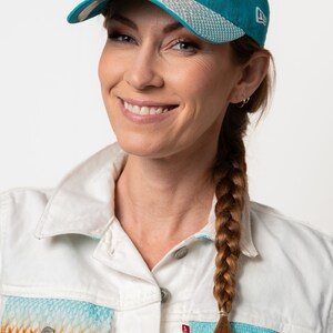 K&ouml;nnte beinhalten: Eine Frau mit langen braunen Haaren, die zu einem Zopf geflochten sind, tr&auml;gt eine wei&szlig;e Jeansjacke mit einer Tasche in einem t&uuml;rkisfarbenen und orangefarbenen Muster. Sie tr&auml;gt auch eine t&uuml;rkisfarbene Baseballkappe mit einem wei&szlig;en gestickten Logo eines Delfins und einer Sonne.