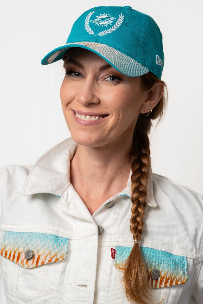 K&ouml;nnte beinhalten: Eine Frau mit langen braunen Haaren, die zu einem Zopf geflochten sind, tr&auml;gt eine wei&szlig;e Jeansjacke mit einer Tasche in einem t&uuml;rkisfarbenen und orangefarbenen Muster. Sie tr&auml;gt auch eine t&uuml;rkisfarbene Baseballkappe mit einem wei&szlig;en gestickten Logo eines Delfins und einer Sonne.