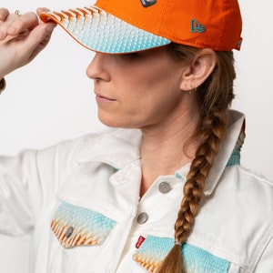 Może przedstawiać: Kobieta w białej dżinsowej kurtce z niebiesko-pomarańczowym wzorem. Ma na sobie jasno pomarańczową czapkę bejsbolową z logo Miami Dolphins i logo New Era. Czapka ma niebieski i biały wzorzysty daszek.