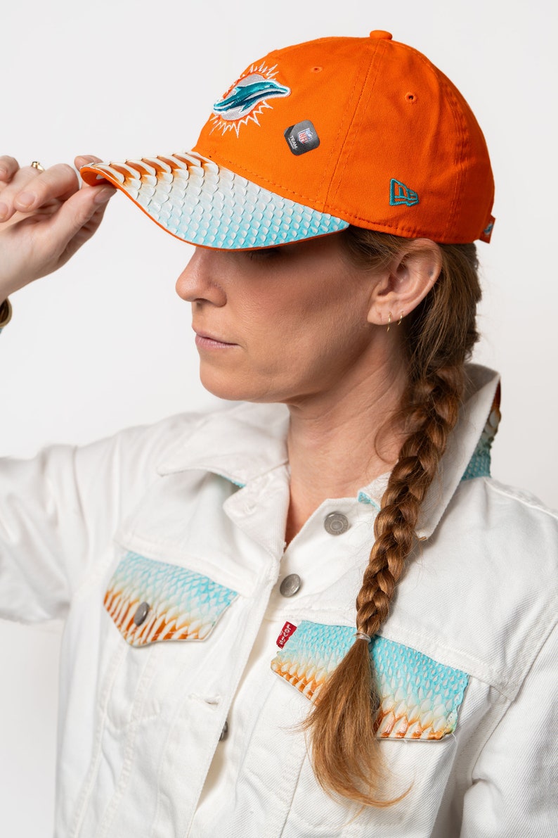 K&ouml;nnte beinhalten: Eine Frau tr&auml;gt eine wei&szlig;e Jeansjacke mit einem blauen und orangenen Muster. Sie tr&auml;gt eine leuchtend orangefarbene Baseballkappe mit einem Miami Dolphins Logo und einem New Era Logo. Die Kappe hat einen blauen und wei&szlig;en gemusterten Schirm.