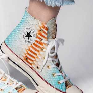 Puede incluir: Zapatillas Converse de caña alta con base blanca y un patrón de piel de serpiente azul, naranja y blanco. Las zapatillas tienen una suela de goma blanca y un logotipo Converse negro con el texto "Chuck Taylor" y "All Star".