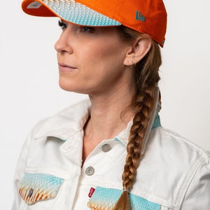 K&ouml;nnte beinhalten: Eine Frau tr&auml;gt eine wei&szlig;e Jeansjacke mit einem blauen und orangefarbenen Muster. Sie tr&auml;gt eine leuchtend orangefarbene Baseballkappe mit einem t&uuml;rkisfarbenen Delfinlogo und dem Schriftzug "Miami Dolphins" auf der Vorderseite. Die Kappe hat einen wei&szlig;en und blauen gemusterten Schirm.