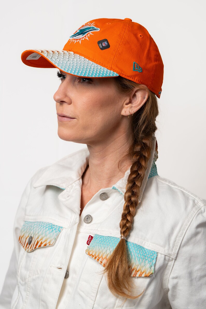 K&ouml;nnte beinhalten: Eine Frau tr&auml;gt eine wei&szlig;e Jeansjacke mit einem blauen und orangefarbenen Muster. Sie tr&auml;gt eine leuchtend orangefarbene Baseballkappe mit einem t&uuml;rkisfarbenen Delfinlogo und dem Schriftzug "Miami Dolphins" auf der Vorderseite. Die Kappe hat einen wei&szlig;en und blauen gemusterten Schirm.