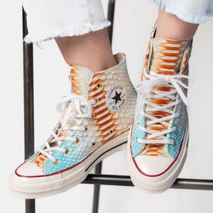 Pu&ograve; includere: Un paio di scarpe Converse alte con base bianca e un motivo a pelle di serpente in tonalit&agrave; di blu, arancione e giallo. Il logo Converse e il testo "All Star" sono visibili sul lato delle scarpe.