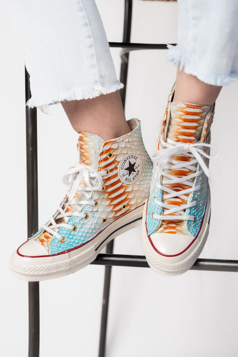 Pu&ograve; includere: Un paio di scarpe Converse alte con base bianca e un motivo a pelle di serpente in tonalit&agrave; di blu, arancione e giallo. Il logo Converse e il testo "All Star" sono visibili sul lato delle scarpe.
