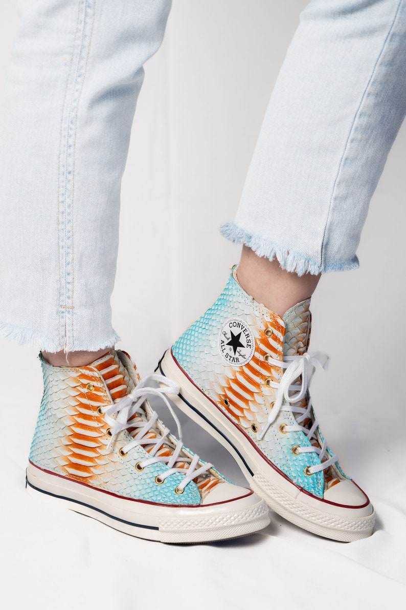 Pu&ograve; includere: Sneakers Converse alte con un motivo a stampa di pelle di serpente blu, arancione e bianco. Le scarpe hanno una suola in gomma bianca e lacci neri. Il logo Converse All Star &egrave; visibile sul lato della scarpa.