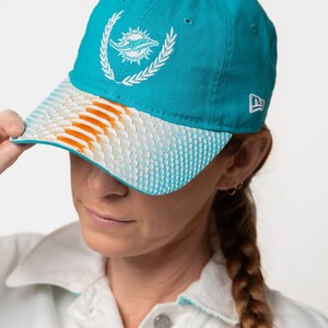 K&ouml;nnte beinhalten: Eine t&uuml;rkisblaue Baseballkappe mit einem wei&szlig;en gestickten Delfinlogo und einem wei&szlig;en und orangefarbenen gemusterten Schirm. Die Kappe hat ein New Era Logo an der Seite.