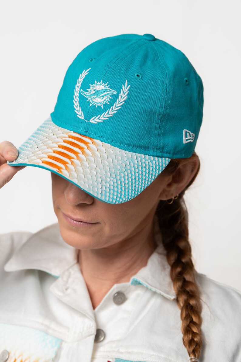 K&ouml;nnte beinhalten: Eine t&uuml;rkisblaue Baseballkappe mit einem wei&szlig;en gestickten Delfinlogo und einem wei&szlig;en und orangefarbenen gemusterten Schirm. Die Kappe hat ein New Era Logo an der Seite.