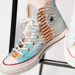 Pu&ograve; includere: Sneakers Converse bianche alte con un motivo a pelle di serpente blu e arancione. Le sneakers hanno una suola in gomma bianca e un logo a stella nero con il testo "Converse All Star" sul lato.