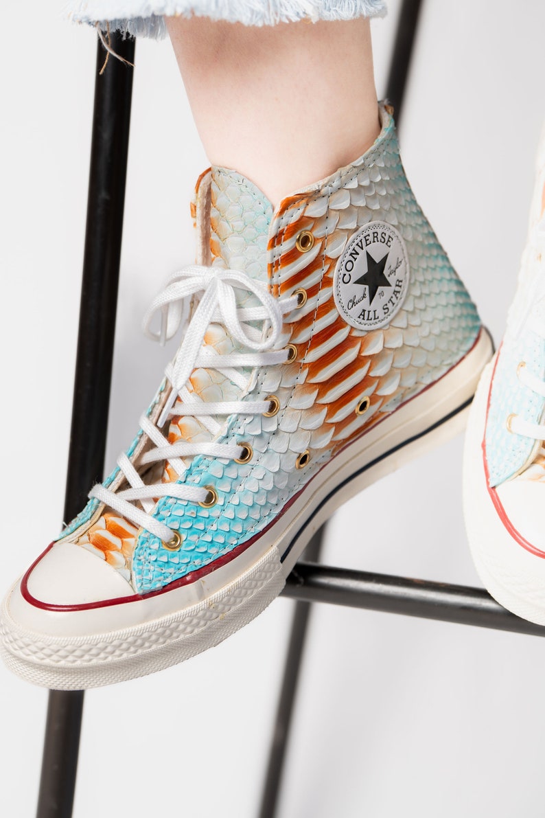 Pu&ograve; includere: Sneakers Converse bianche alte con un motivo a pelle di serpente blu e arancione. Le sneakers hanno una suola in gomma bianca e un logo a stella nero con il testo "Converse All Star" sul lato.