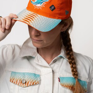 K&ouml;nnte beinhalten: Eine leuchtend orangefarbene Baseballkappe mit einem blauen Delfin-Logo und einer wei&szlig;en und blauen Unterseite mit Muster. Die Kappe hat ein New Era-Logo an der Seite.