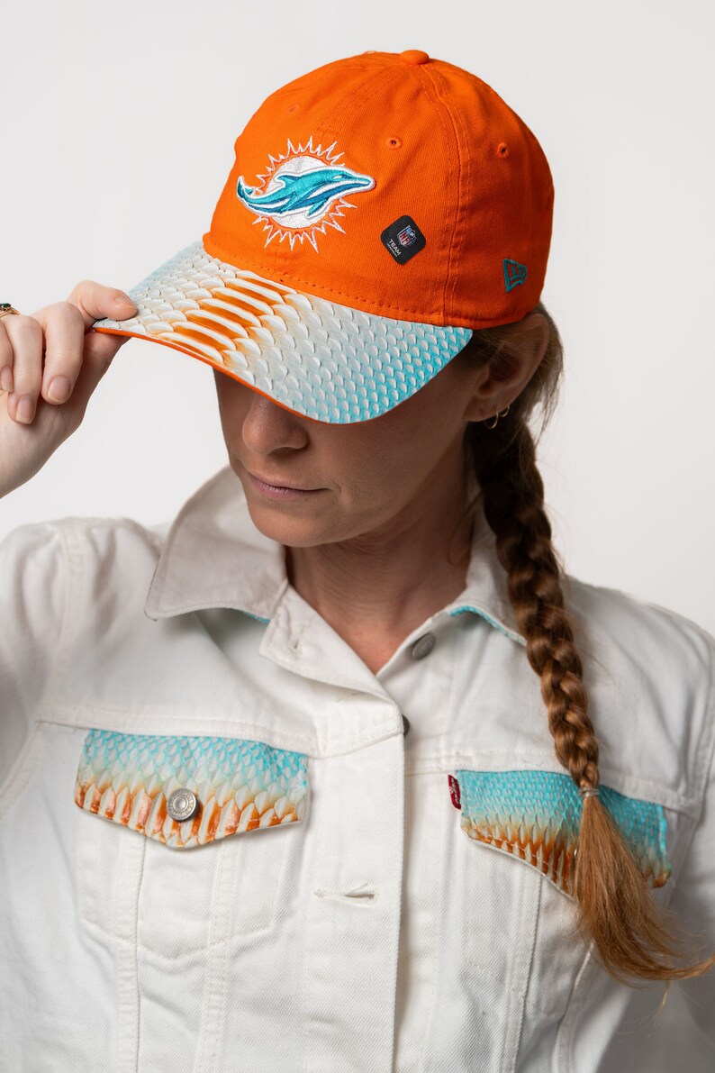 K&ouml;nnte beinhalten: Eine leuchtend orangefarbene Baseballkappe mit einem blauen Delfin-Logo und einer wei&szlig;en und blauen Unterseite mit Muster. Die Kappe hat ein New Era-Logo an der Seite.