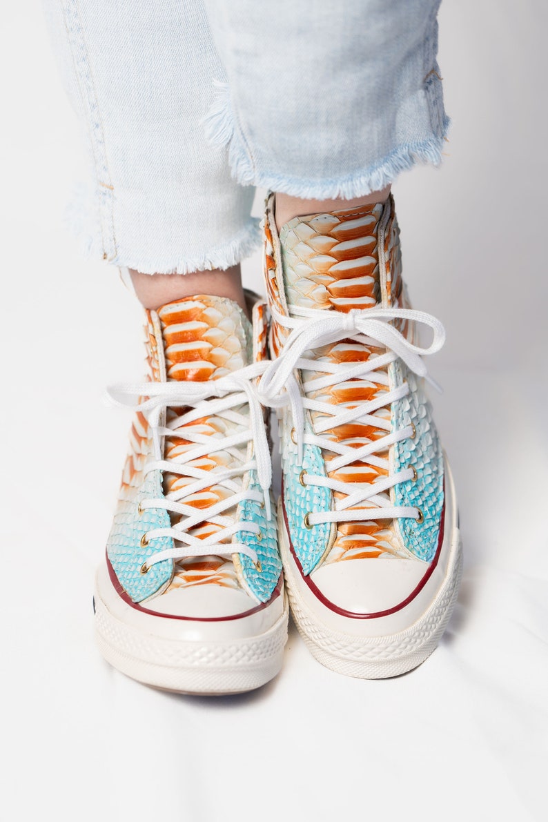 Pu&ograve; includere: Un paio di sneakers bianche alte con tessuto a fantasia di pelle di serpente arancione e blu. Le sneakers hanno lacci bianchi e una suola in gomma bianca.