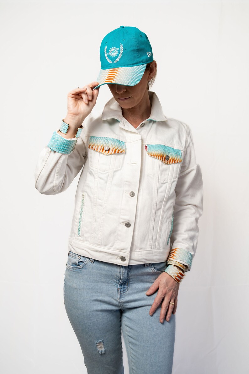 K&ouml;nnte beinhalten: Eine wei&szlig;e Jeansjacke mit farbenfrohem, gemustertem Stoff an den &Auml;rmelb&uuml;ndchen und Taschen. Die Jacke wird &uuml;ber einer hellblauen Jeans getragen. Eine blaugr&uuml;ne Baseballkappe mit wei&szlig;em Logo wird auf dem Kopf getragen.