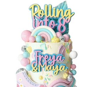 Op de afbeelding: Een versierde taart met de woorden "Rolling into 8" en de namen "Freya & Maya". De taart is versierd met pastelkleurige decoraties, waaronder een regenboog, een rolschaats en discoballen. Roze, blauwe en gele accenten worden gebruikt.