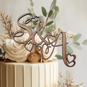 Könnte beinhalten: Eine zweistöckige Hochzeitstorte mit weißem Zuckerguss und einem Kuchenaufsatz aus Holz. Der Aufsatz zeigt die Buchstaben "S & A" verschlungen mit Herzen. Die Torte ist mit weißen Rosen, Trockenblumen und Grünzeug verziert.