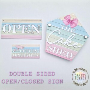 Op de afbeelding: Een set pastelkleurige houten borden. Eén bord leest "OPEN", een ander zegt "CCTV IN OPERATION", en een derde, in de vorm van een huis, zegt "THE Cake SHED" met een roze strik. De borden zijn dubbelzijdig.