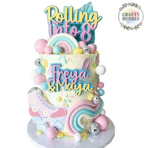 May include: A celebratory cake decorated with the words "Rolling into 8" and the names "Freya & Maya". The cake features pastel decorations, including a rainbow, roller skate, and disco balls. The cake is presented on a white cake stand.
