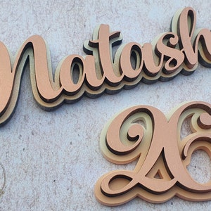 Könnte beinhalten: Ein mehrschichtiges Holz-Namensschild mit dem Namen "Natasha" und der Zahl "26" in einer kursivschriftlichen Schriftart. Das Schild ist in Kupferfarbe mit einem hellbraunen Umriss bemalt.
