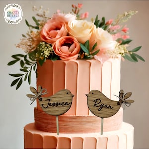 May include: A three-tiered wedding cake with peach-coloured frosting and floral decorations. Two wooden bird cake toppers are featured, one with the name "Jessica" and the other with "Ryan". The cake is topped with peach and cream roses and greenery.
