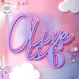 Puede incluir: Un adorno para tarta en capas en rosa y lavanda, con el nombre "Olive" y el número "6" en una fuente cursiva. El adorno está sobre un fondo rosa y morado pastel, con formas de nubes blancas y perlas decorativas.