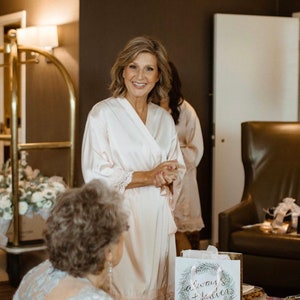 May include: A group of women in a hotel room getting ready for a wedding. One woman is wearing a white robe with lace trim and is smiling at the camera. Another woman is standing behind her, also wearing a white robe. A gift bag with the text "always + forever" is on a table in the foreground.