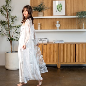 May include: A white lace robe with a satin lining. The robe is being worn by a person standing in a room with a wooden cabinet and a plant.