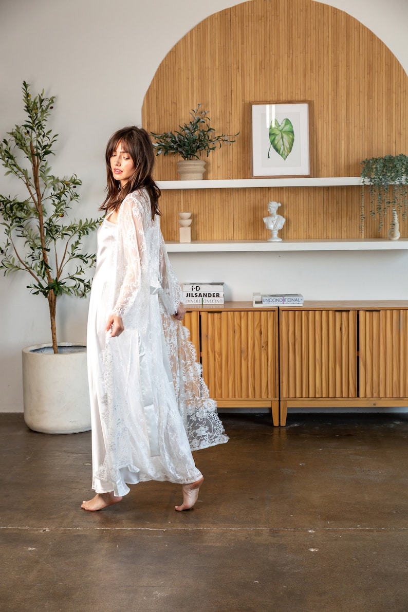 May include: A white lace robe with a satin lining. The robe is being worn by a person standing in a room with a wooden cabinet and a plant.