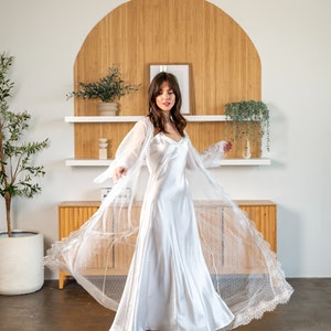 May include: A white satin slip dress with a sheer lace overlay. The dress has a V-neckline and a flowing skirt. The model is wearing the dress in a room with a wooden wall behind her.